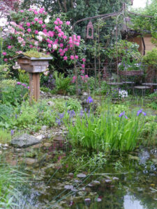 Der Teich mit Rosenrondell, Insektenhotel und Rhododendronsenke in Wurzerlsgarten.